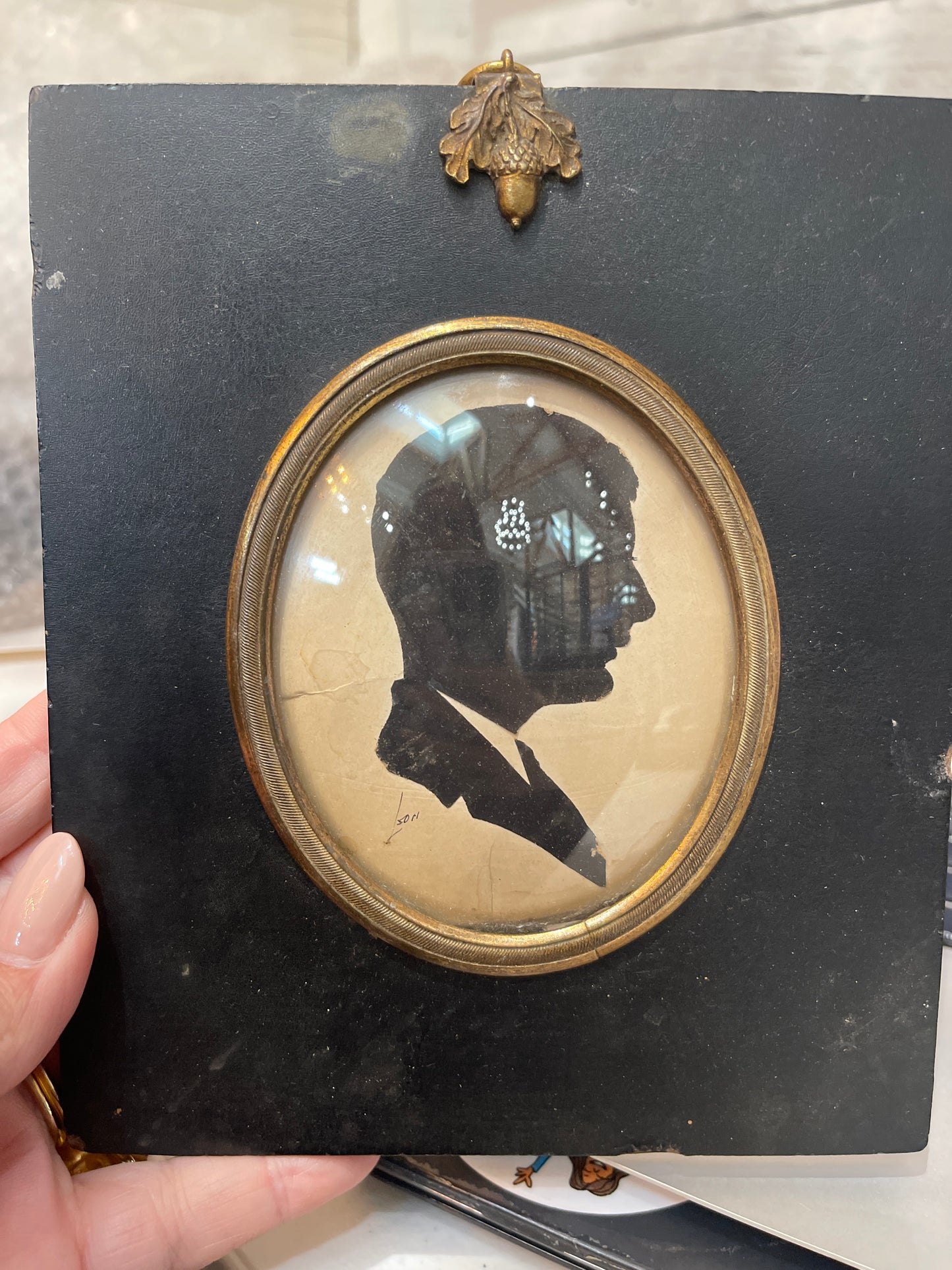 A silhouette miniature portrait of a man in a frame, held by a hand against a black background.