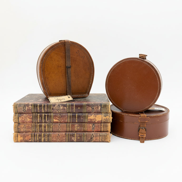 Stack of vintage books and a brown leather hat box on a glass shelf.