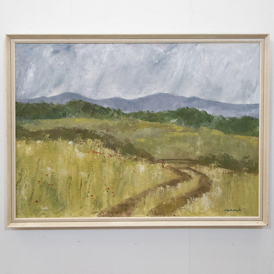 A landscape painting depicting a pathway in a field with a mountain range in the background and a blue sky overhead, framed and hanging on a wall.